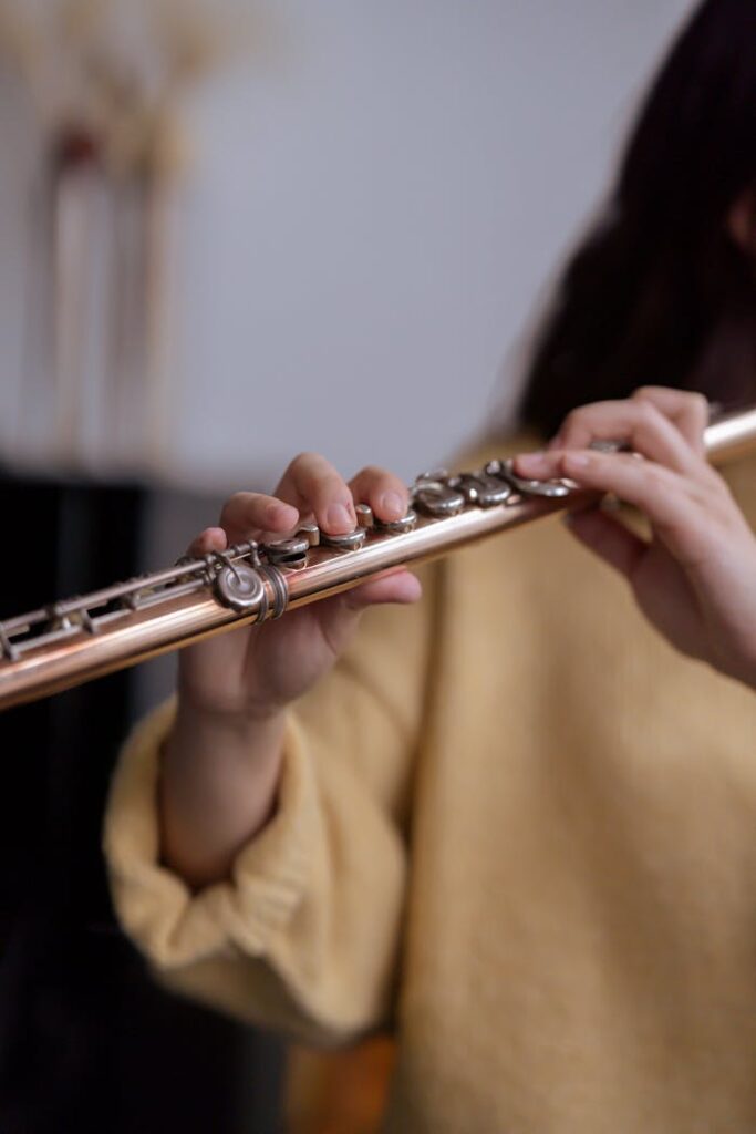 Crop faceless female musician in warm yellow sweater playing on flute in light classroom