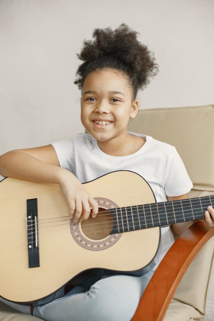 Young girl smiling while playing an acoustic guitar, seated indoors.